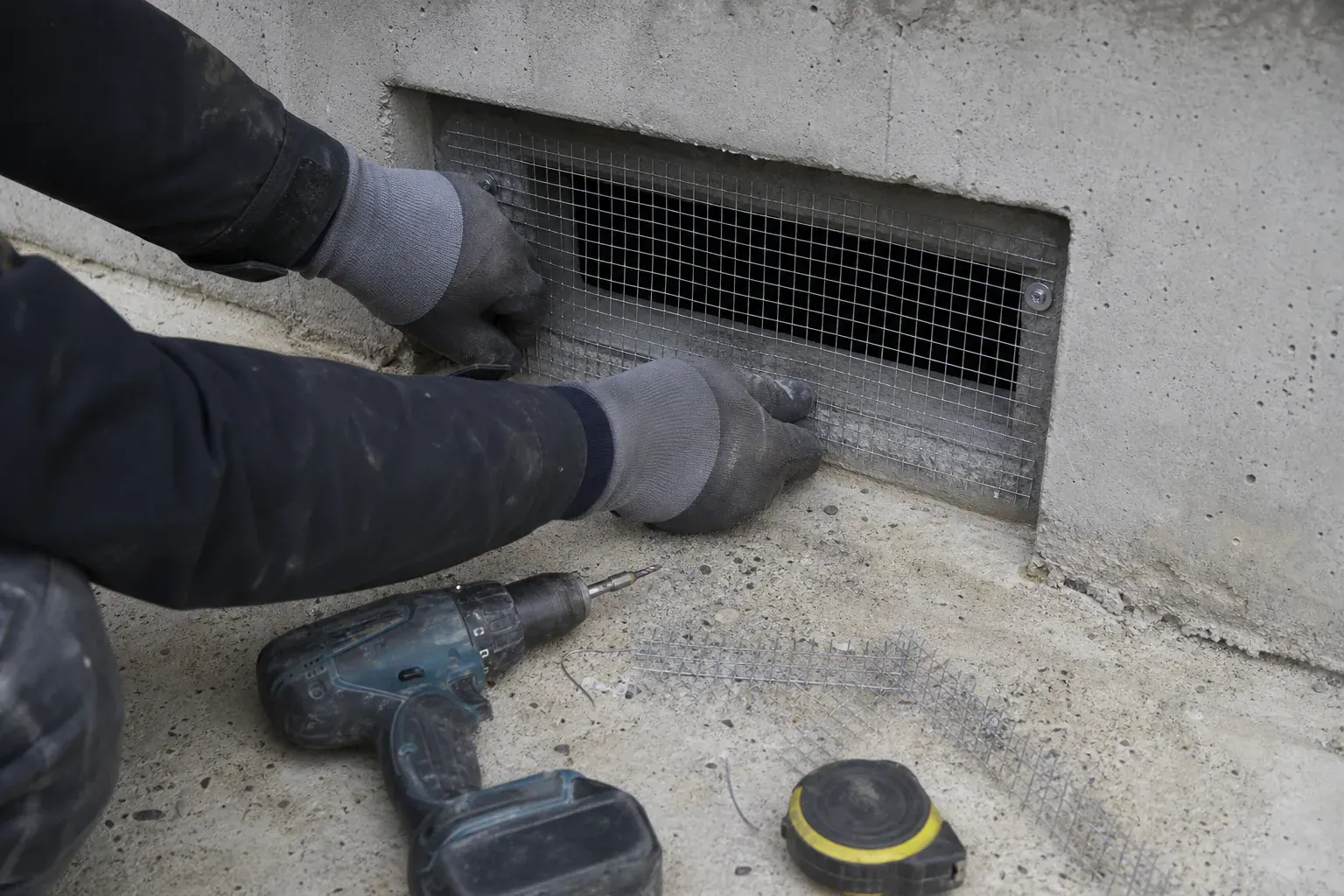 Mains gantées fixant une grille métallique fine sur une ouverture de ventilation basse en mur de béton